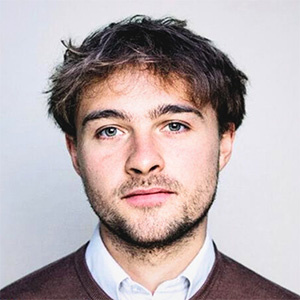 Tom Butler stands in front of a white background. Tom wears a maroon jumper and white shirt.