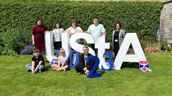 5 people standing behind and 3 people sitting in front of large white letter spelling UStA (spelling University of St Andrews)