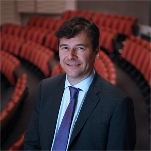 Professor Andrew Jones standing in a lecture theatre in front of rows of red chairs. Professor Jones wears a blue tie and black suit jacket.