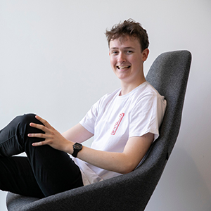 Jamie Bankhead smiling and sitting in a modern grey chair. He wears a white t-shirt and black trousers.