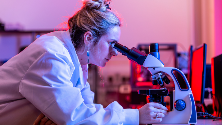 A woman wearing a lab coat looks through a microscope in a lab