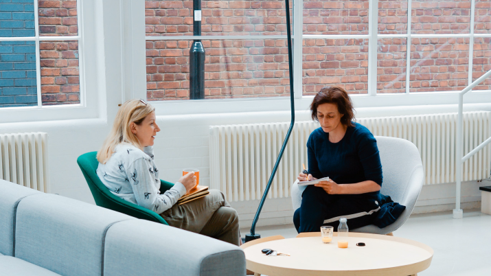 Two colleagues sit on a sofa in a light room discussing something.