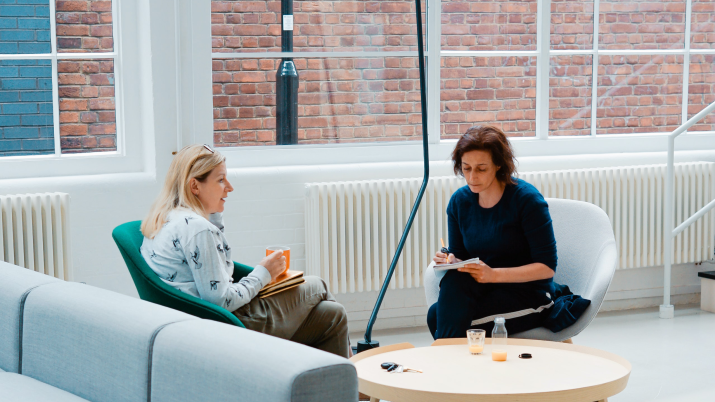 Two colleagues sit on a sofa in a light room discussing something. 