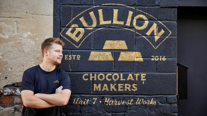 Max Bullion standing next to a wall with the Bullion Chocolate logo. It reads: 'Bullion established 2016. Chocolate makers. Unit 7, Harvest Works'