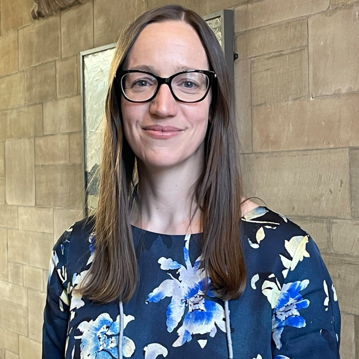 Dr Nia Jones standing against a brick wall. Dr Jones wears a floral blue top and glasses