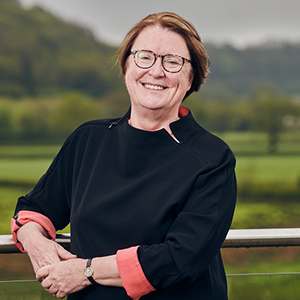 Professor Elwen Evans stands outside. In the background, there is a view of the Welsh countryside, with green fields and trees.