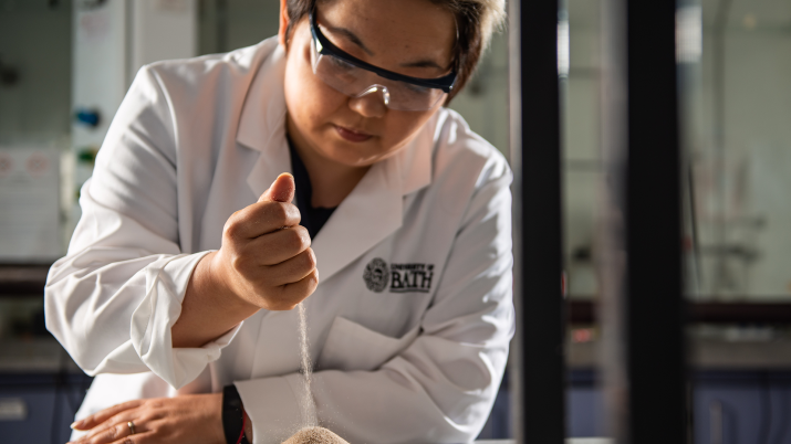Dr Asel Sartbaeva in a lab wearing a white lab coat and goggles. She is pouring sand into a pile.
