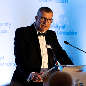 Professor Graham Baldwin making a speech. He wears black tie and stands in front of a backdrop with the University of Central Lancashire logo.