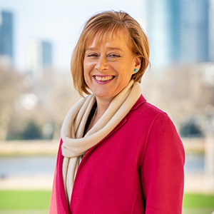 Jane Harrington smiling against a backdrop of the view of London's skyscrapers from Greenwich. She is wearing a pink wool coat and white scarf.