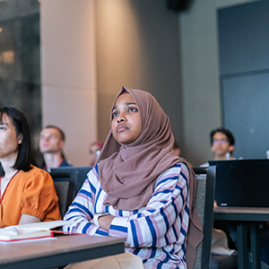 Maisha Saida sitting in a classroom looking to the front with interest. She wears a hijab and an outfit with a striped pattern.