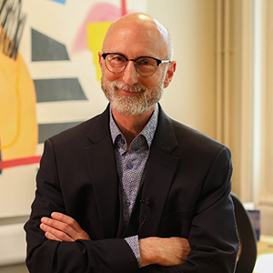 Nic Beech smiling with his arms crossed in front of a wall where a piece of modern art is hanging. He is wearing a suit with no tie.