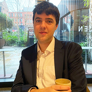 Rowan sitting in a café holding a coffee. He is wearing a white shirt and a black blazer.
