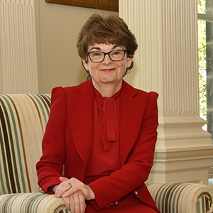 Sally Mapstone smiling while sitting on a striped armchair in a red blouse and red blazer.