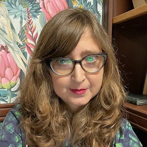Sandra sitting in front of a bookshelf and a wall with floral wallpaper.