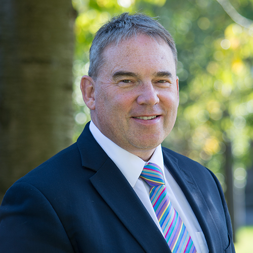 Headshot of Professor Sir Steve West. He's standing outside in a park with a leafy background. He wears a shirt and stripy tie with a dark jacket.