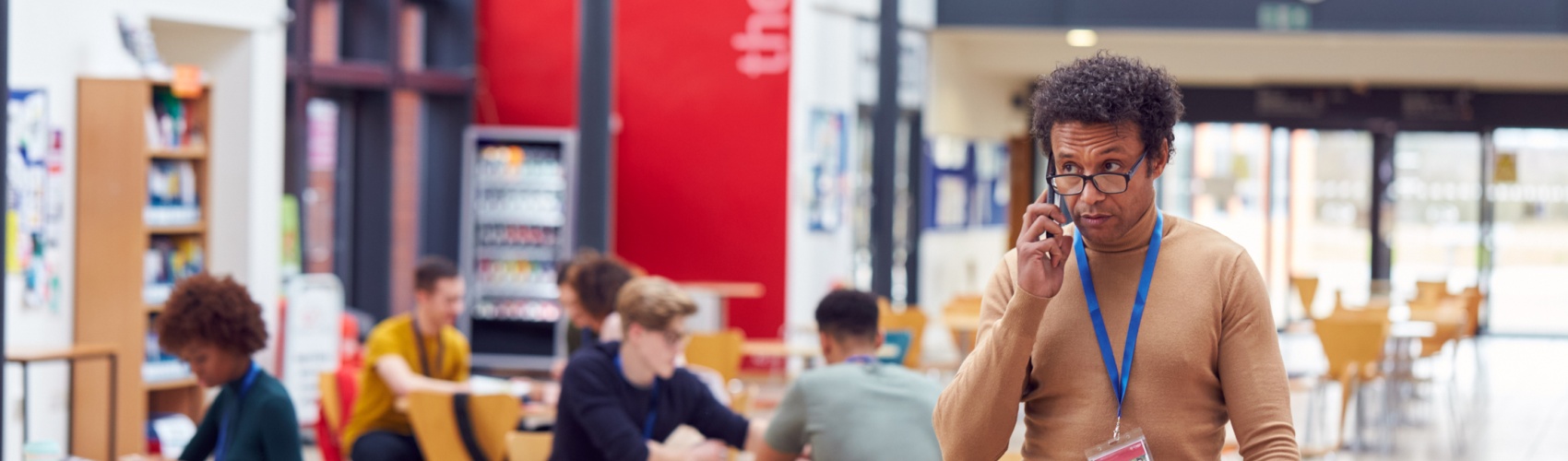 Communal Area Of Busy College Campus With Students Working At Tables And Tutor On Phone