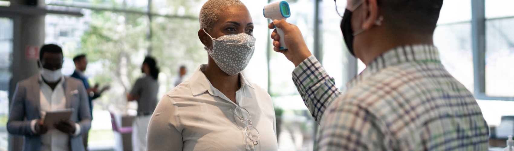 Receptionist measuring temperature of female employee at office's entrance - with face mask