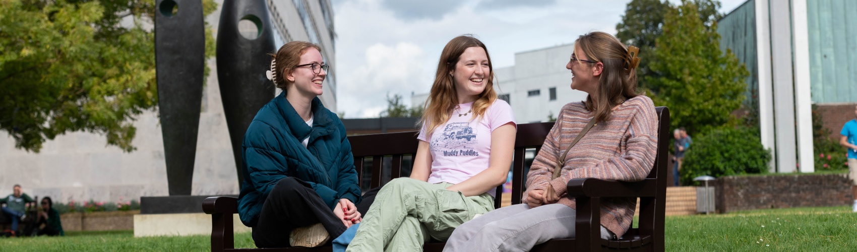 Three students relax and chat on a bench on a leafy university campus. There is a modern sculpture in the background.