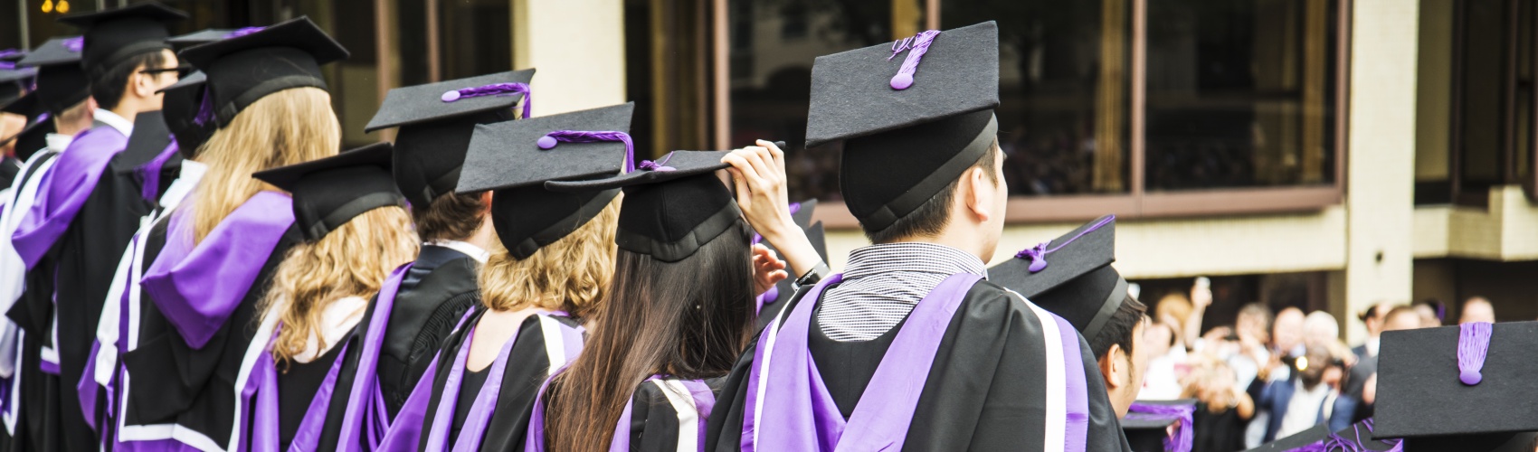 Back view of a university graduation ceremony. The graduates wear black and purple gowns and graduation caps.