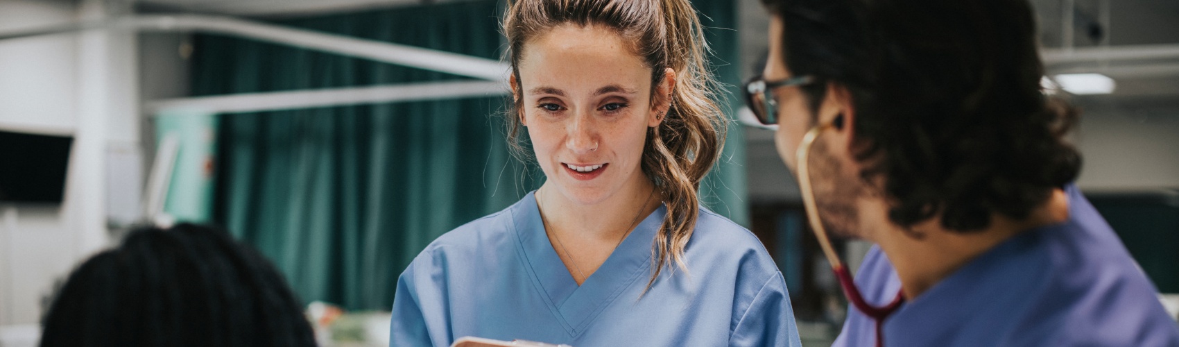 A male doctor examining a patient with a stethoscope. The female doctor writes notes on a clipboard.