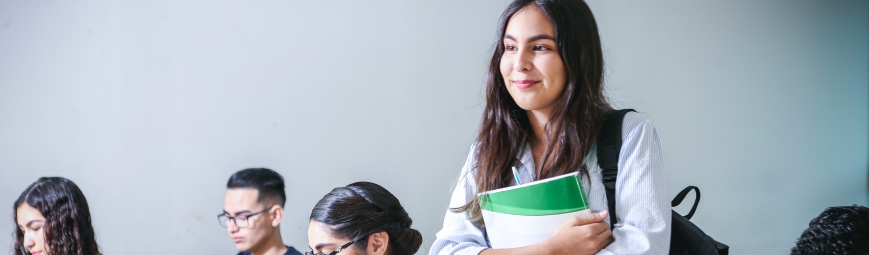 Students in a classroom, one in the forefront standing and smiling with a textbook