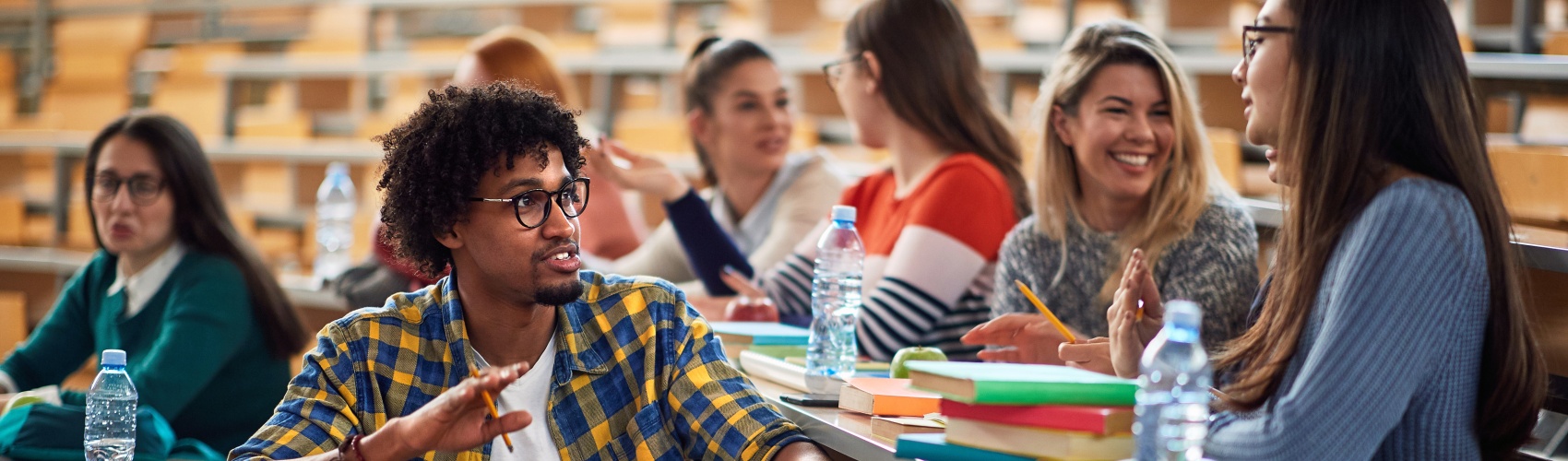 A group of students sat in a lecture hall chatting, smiling and studying. 