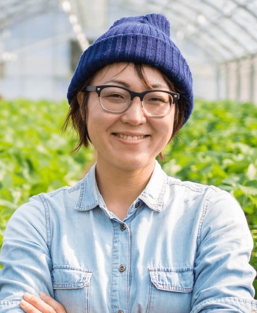 Woman wearing blue bobble hat inside a greenhouse with plants behind her.