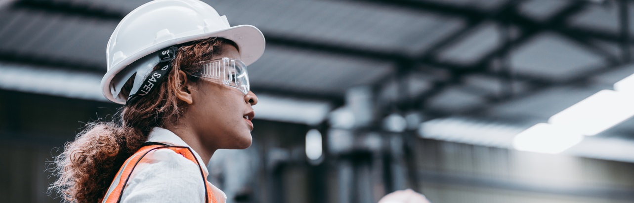 Female industrial engineer wearing a white helmet while standing in a heavy industrial factory behind she talking with workers, Various metal parts of the project
