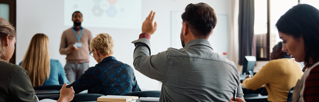 A classroom with students seated in rows of long desks and a teacher standing at the front with a presentation displayed on the wall behind him. One student in the middle raises their hand to ask a question.