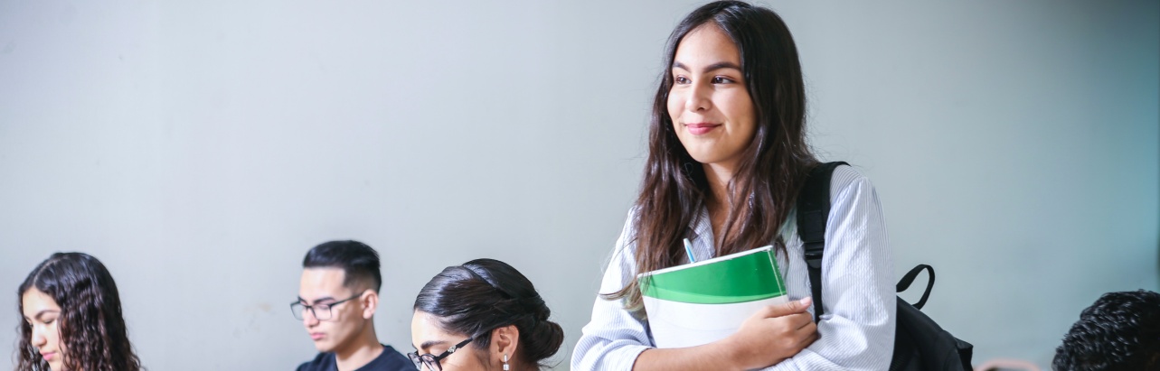 Students in a classroom, one in the forefront standing and smiling with a textbook