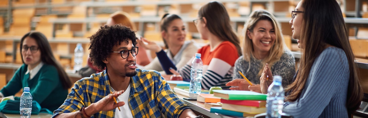 A group of students sat in a lecture hall chatting, smiling and studying. 