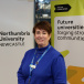 Roisin Currie, Greggs Cheif Executive stands in front of a Northumbria University and Universities UK banner. Roisin wears a blue top and Greggs lanyard.