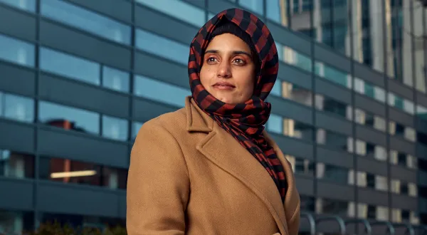 Dr Beenish Siddique standing in front of a modern university building on a sunny day. She wears a red and black patterned hijab and a light brown wool coat.