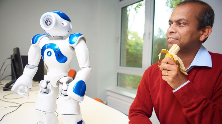 Professor Ardhendu Behera sitting next to a robot while eating a banana